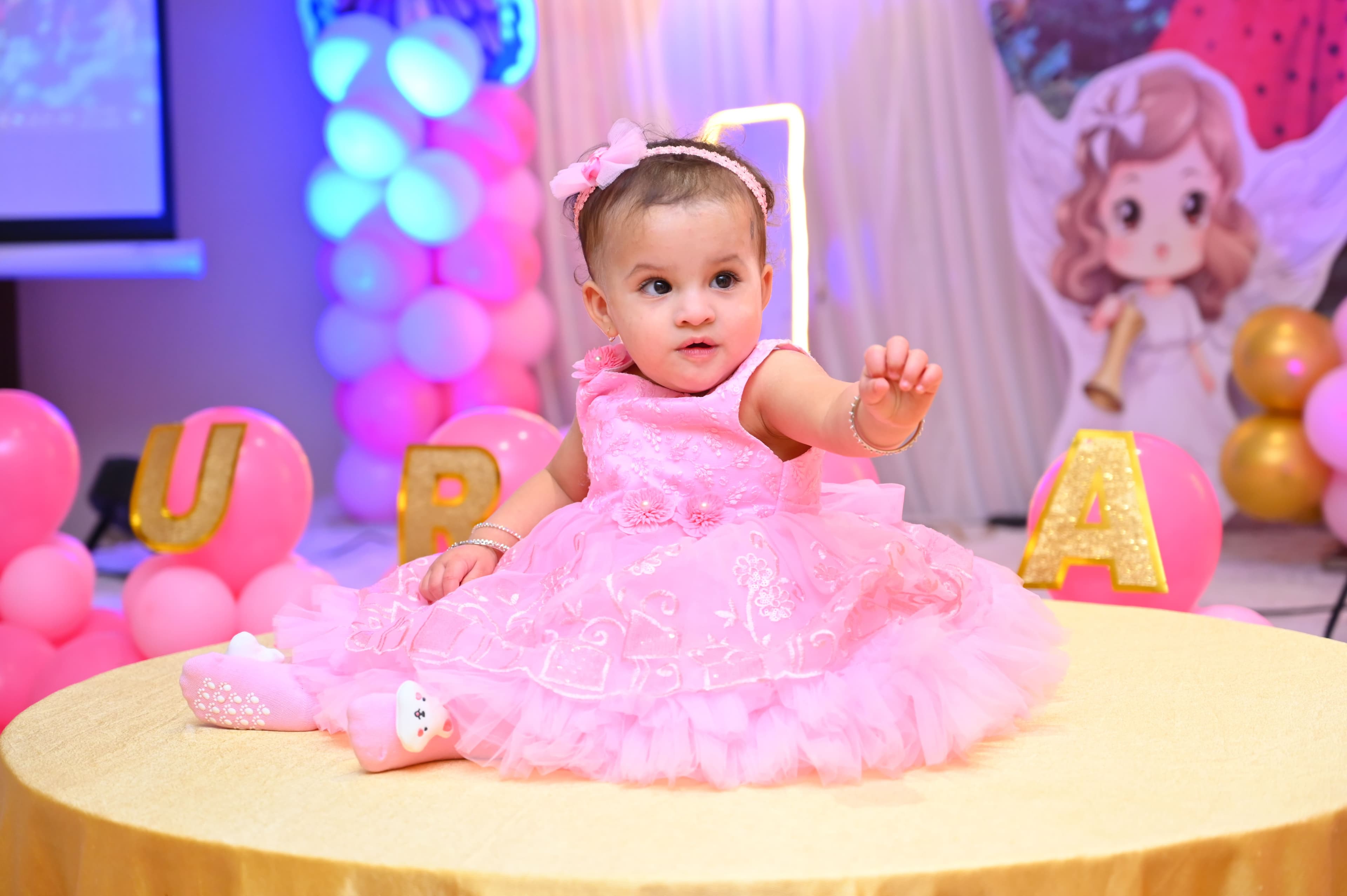 Baby girl in a pink dress reaching out on a gold-draped table, first birthday