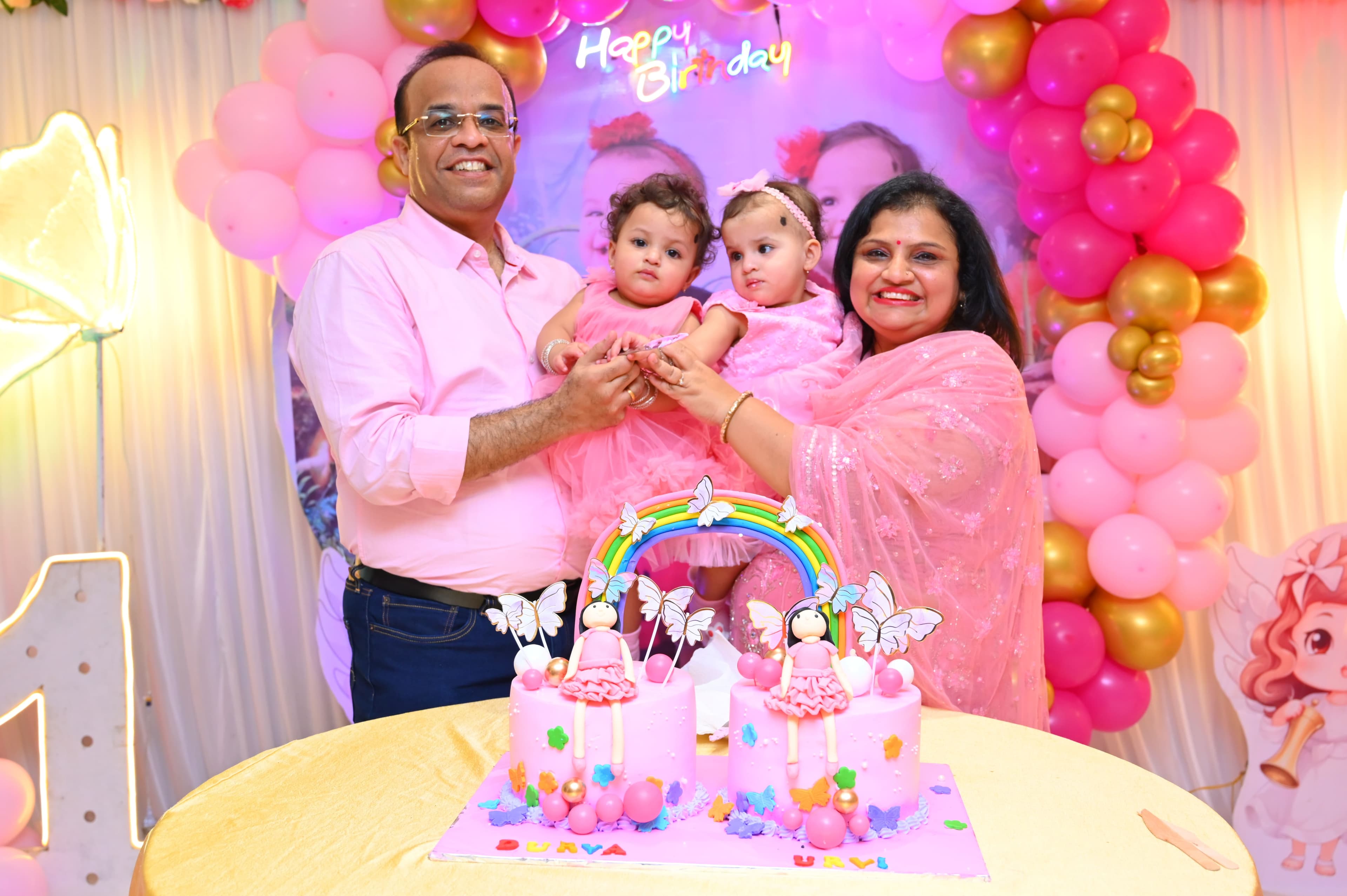 Family with two baby girls behind a rainbow birthday cake