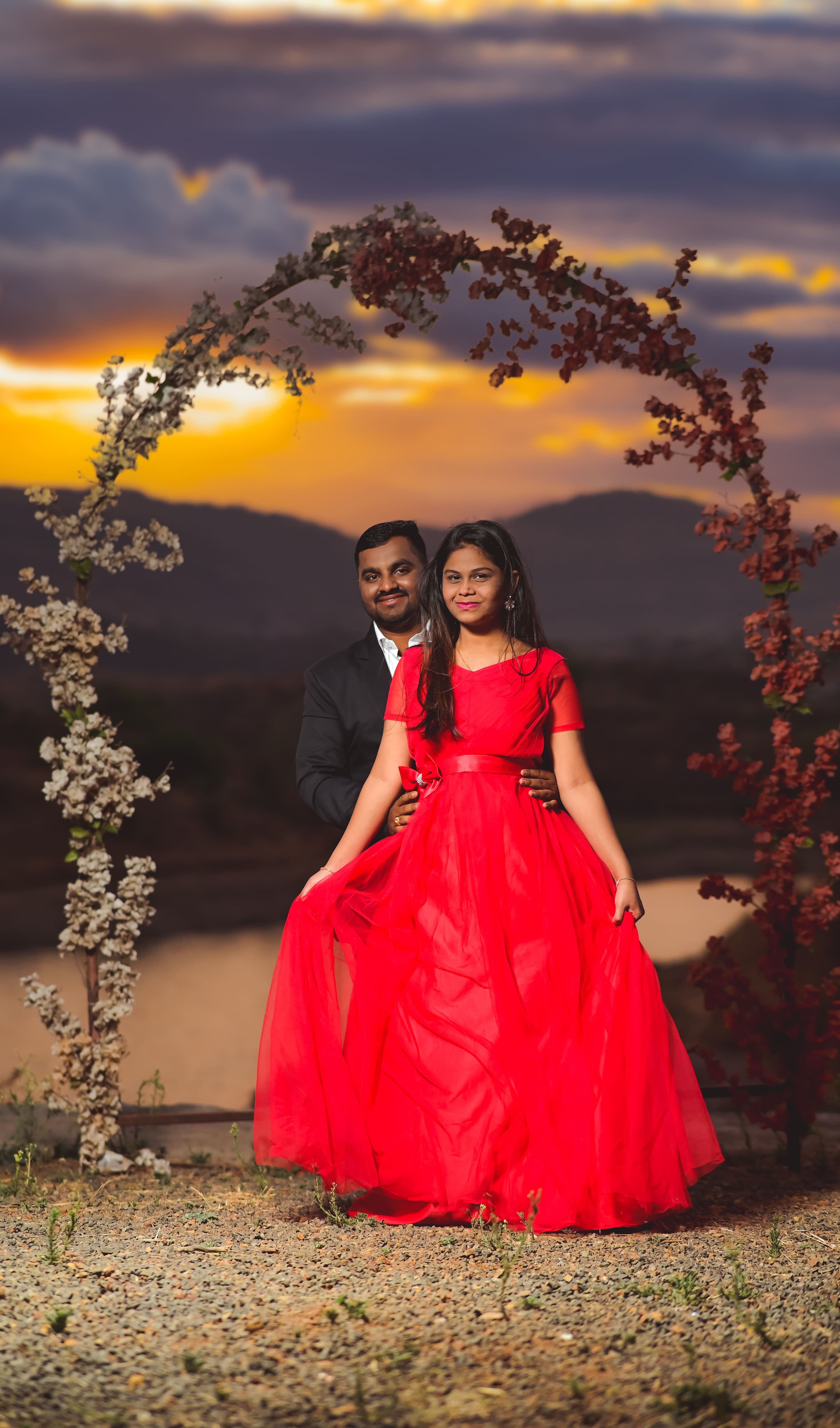 Bride-to-be in a red gown with her partner under a floral arch at sunset