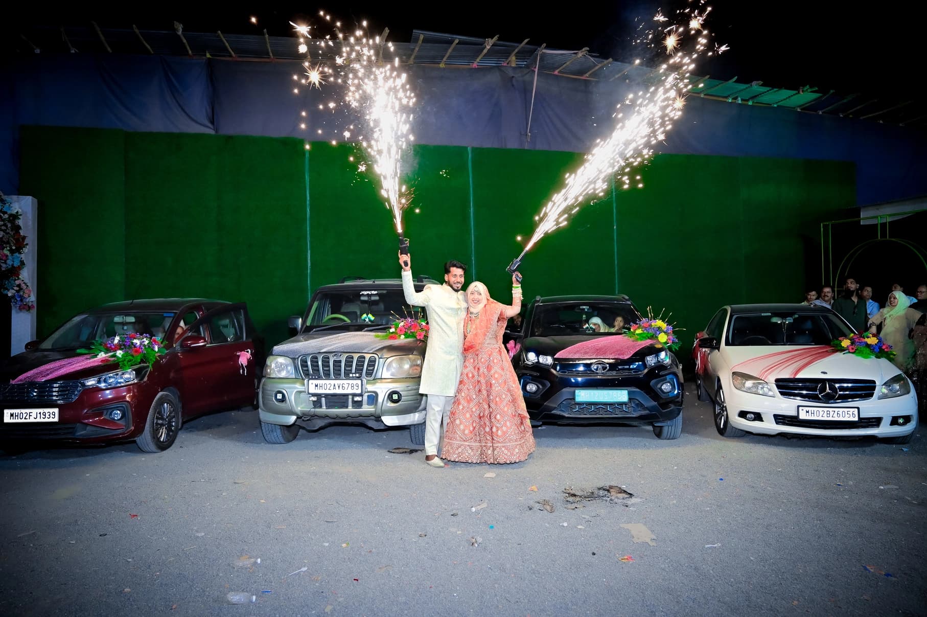 Bride and groom with cold-pyro fireworks beside decorated wedding cars at night
